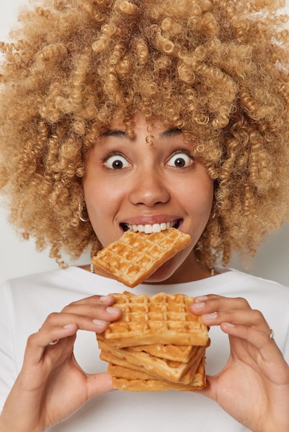 Imagem vertical de mulher de cabelo encaracolado surpresa come delicioso waffle belga caseiro doce gosta de sobremesa favorita tem olhos arregalados poses contra fundo branco comida de altas calorias quebra de dieta