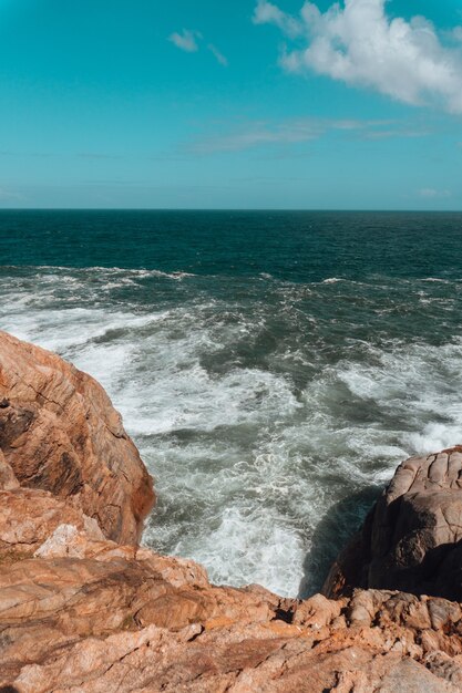 Imagem vertical de rochas cercadas pelo mar sob um céu azul e luz do sol