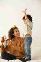 Foto grátis imagem vertical de um menino egocêntrico de dois anos exigindo de sua linda jovem mãe que lhe desse seu brinquedo de pelúcia. mãe mestiça sentada na cama branca brincando com o filho pequeno