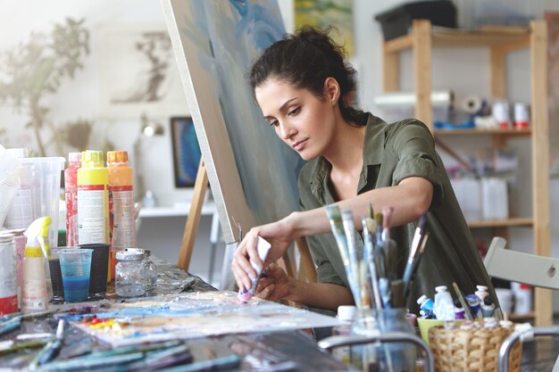 Imagens de sério concentrada jovem artista feminina caucasiana, sentado na mesa com acessórios de pintura, segurando o tubo de tinta a óleo, misturando cores na paleta; pintura inacabada sobre tela perto dela