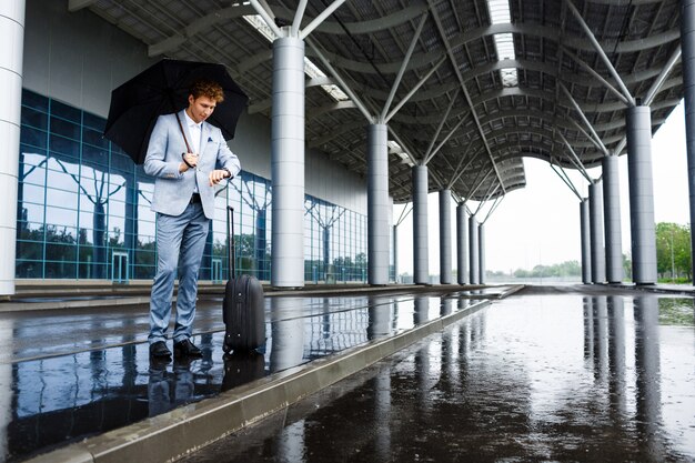 Imagens do jovem empresário ruivo segurando guarda-chuva preta na chuva e olhando para o relógio na estação