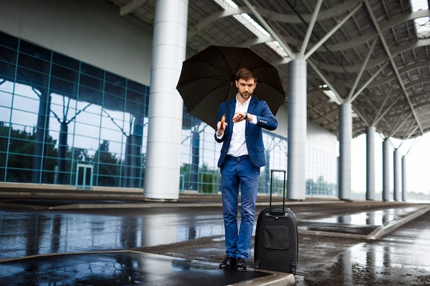 Imagens do jovem empresário segurando a mala e o guarda-chuva, olhando no relógio, esperando na estação chuvosa