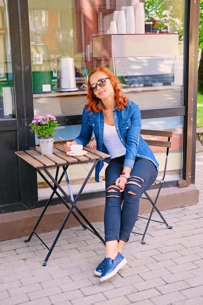 Imgae de corpo inteiro de mulher ruiva bebe café à mesa em um café na rua.