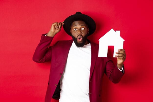 Imobiliária. Homem negro alegre mostrando a casa de papel e sorrindo, recomendando o corretor, em pé sobre um fundo vermelho