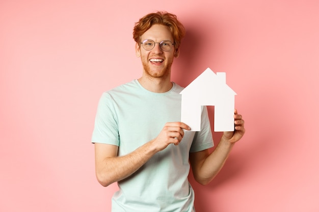 Imobiliária. Homem ruivo bonito de camiseta e óculos, mostrando o recorte da casa de papel e sorrindo, em pé sobre um fundo rosa.