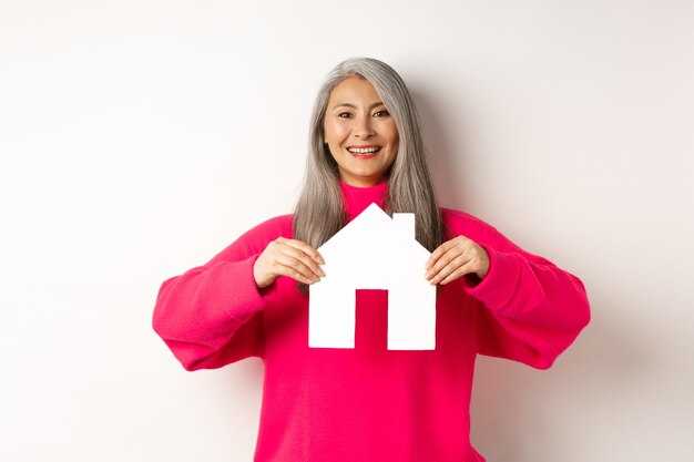 Imobiliária linda avó asiática mostrando a casa de papel e sorrindo feliz em pé de suor rosa ...