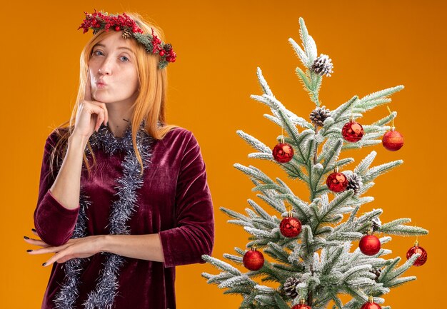 Impressionada, franzindo os lábios, jovem linda garota em pé perto da árvore de Natal usando um vestido vermelho e grinalda com uma guirlanda no pescoço colocando o dedo na bochecha