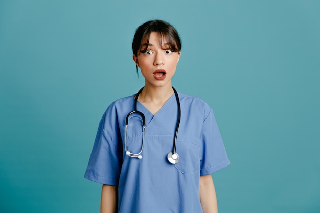 Impressionada jovem médica usando estetoscópio uniforme de quinto isolado em fundo azul