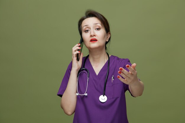 Impressionada médica de meia-idade vestindo uniforme e estetoscópio no pescoço, olhando para o lado, esticando a mão em direção à câmera, falando no telefone isolado no fundo verde-oliva