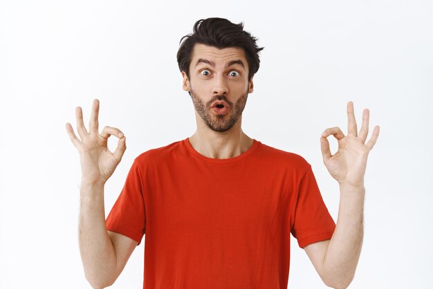 Impressionado animado homem caucasiano bonito com barba em camiseta vermelha mostrando o gesto ok, julgando algo incrível, diga uau, produto maravilhoso em pé, espantado com fundo branco