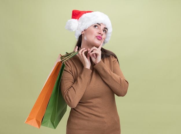 Impressionado ao olhar para uma jovem linda com chapéu de natal segurando uma sacola de presente no ombro isolada em fundo verde oliva