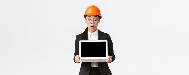 Impressionado arquiteto de engenharia de construção feminino asiático ou gerente na empresa mostrando a tela do laptop com expressão espantada olhando para o diagrama de lucro durante a reunião usar capacete de segurança na fábrica