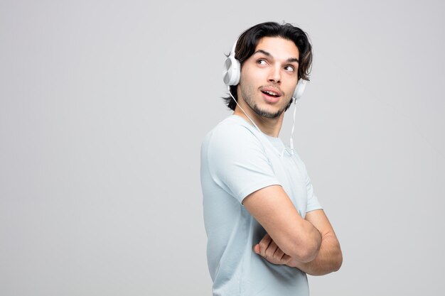 impressionado jovem bonito usando fones de ouvido em vista de perfil com os braços cruzados isolados no fundo branco com espaço de cópia