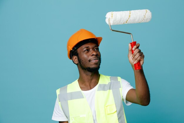 Impressionado jovem construtor americano africano em uniforme segurando escova de rolo isolada em fundo azul