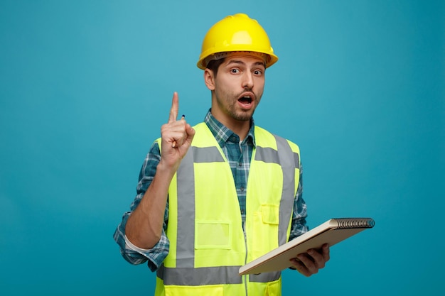 Impressionado jovem engenheiro masculino usando capacete de segurança e uniforme segurando lápis e bloco de notas olhando para câmera apontando para cima isolado em fundo azul