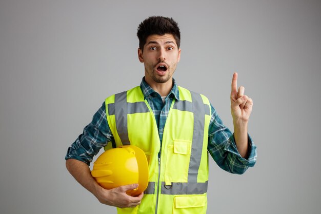 Impressionado jovem engenheiro masculino vestindo uniforme segurando capacete de segurança olhando para câmera apontando para cima isolado no fundo branco