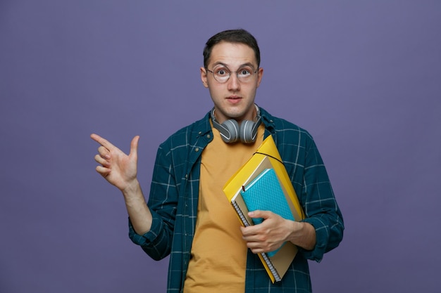 Impressionado jovem estudante do sexo masculino usando fones de ouvido de óculos no pescoço, segurando a pasta do bloco de notas do bloco de notas olhando para a câmera apontando para o lado isolado no fundo roxo