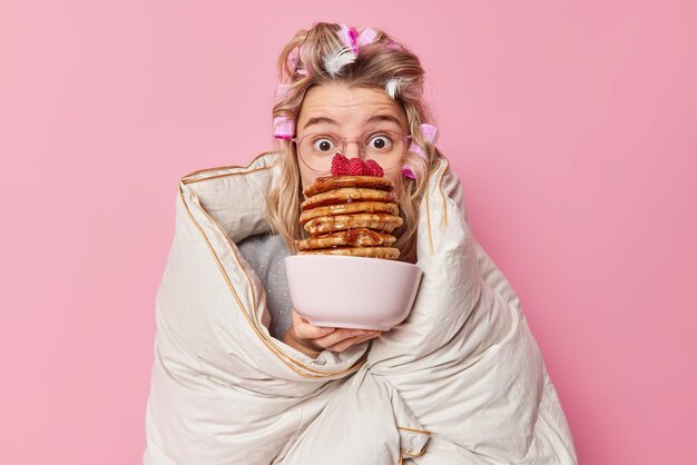 Impressionado jovem europeu com rolos de cabelo embrulhados em edredom detém tigela de panquecas apetitosas com calda usa grandes óculos isolados sobre fundo rosa. Conceito de café da manhã e manhã