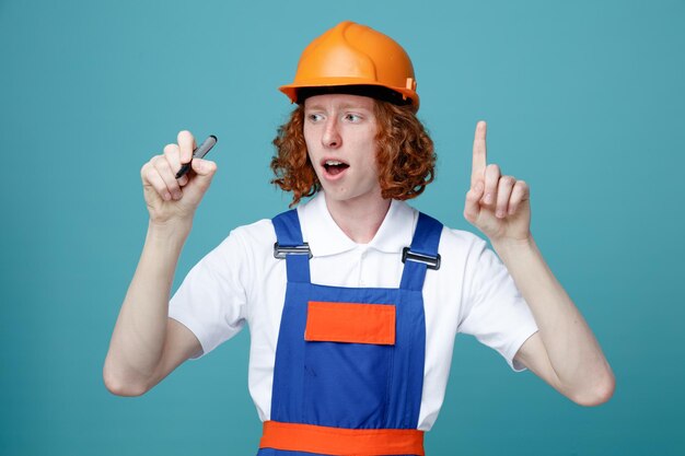 Impressionado mostrando número jovem construtor de uniforme segurando caneta marcador para câmera isolada em fundo azul