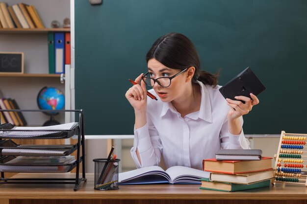 impressionado olhando para o lado jovem professora de óculos segurando calculadora sentado na mesa com ferramentas escolares em sala de aula