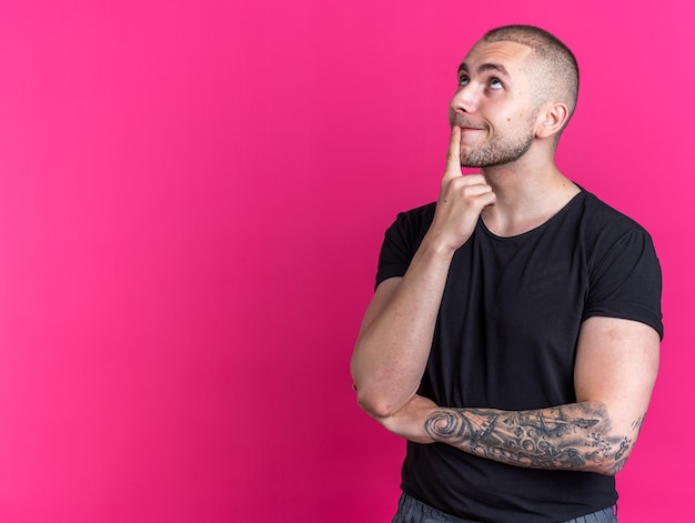 Impressionado olhando para um jovem bonito vestindo camiseta preta e colocando o dedo no queixo isolado em um fundo rosa com espaço de cópia