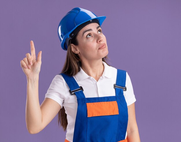 Impressionado olhando para uma jovem construtora de uniforme aponta para cima isolado na parede roxa
