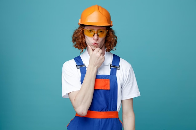 Impressionado queixo agarrado jovem construtor de uniforme isolado em fundo azul