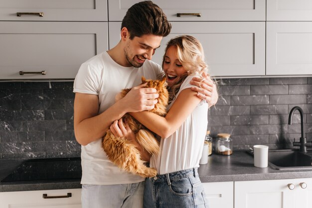 Impressionante garota encaracolada segurando um gato fofo. Retrato interior de casal europeu brincando com seu animal de estimação.