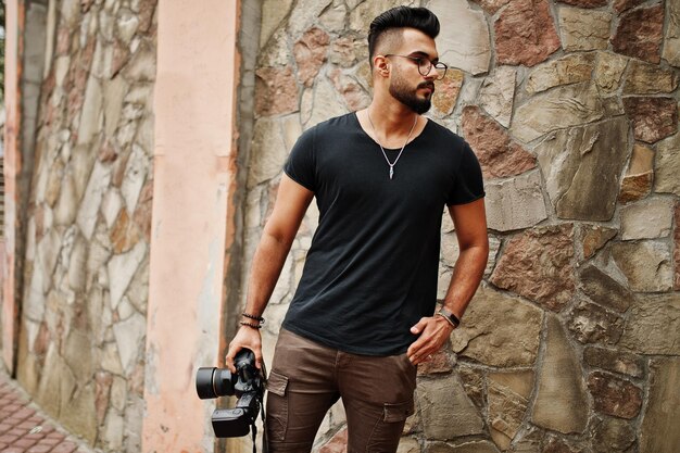 Impressionante lindo e alto fotógrafo macho de barba árabe em óculos e camiseta preta com câmera profissional nas mãos