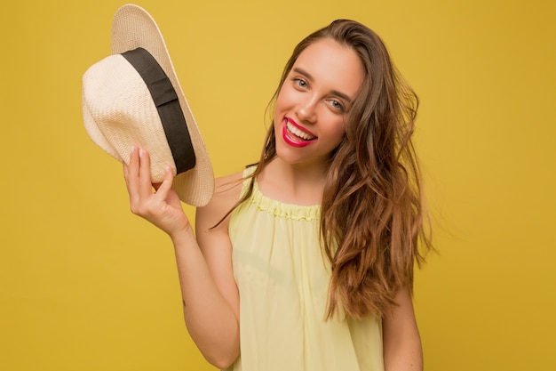 Impressionante modelo feminino de cabelos compridos posando na parede amarela, segurando um chapéu e sorrindo