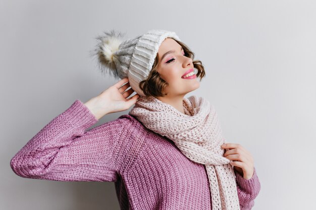 Impressionante mulher caucasiana em suéter roxo posando de brincadeira. Foto interna de menina graciosa usa chapéu de lã branco e cachecol olhando para longe com um sorriso.