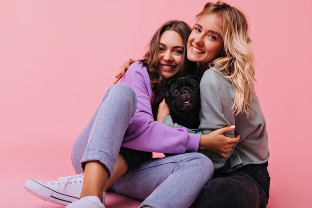 Incríveis irmãs brancas se abraçando durante a sessão de retratos com o cachorro. adoráveis jovens senhoras sentadas na rosa com um lindo bulldog.