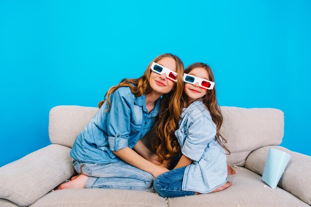 Incrível mãe e filha se abraçando e sorrindo em óculos 3d para a câmera no sofá isolado sobre fundo azul. Expressando verdadeiras emoções felizes, tempo para a família da mãe com o filho fofo, entretenimento juntos