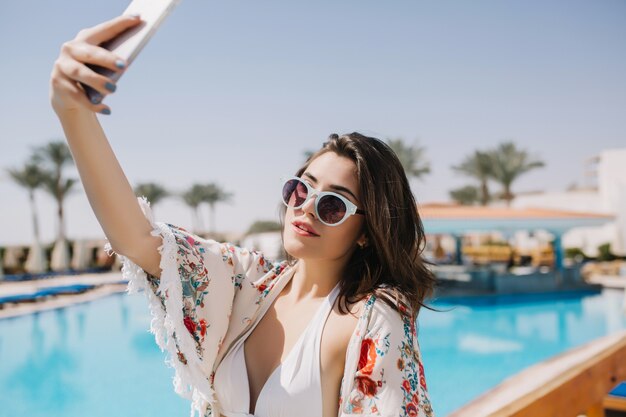 Incrível menina morena de biquíni e camisa da moda, fazendo selfie na piscina ao ar livre na manhã de verão. Retrato de uma jovem encantadora em óculos de sol e maiô branco tirando foto de si mesma