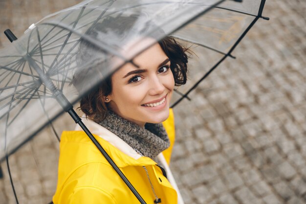 Incrível retrato de jovem de casaco amarelo em pé sob o guarda-chuva transparente com amplo sorriso sincero