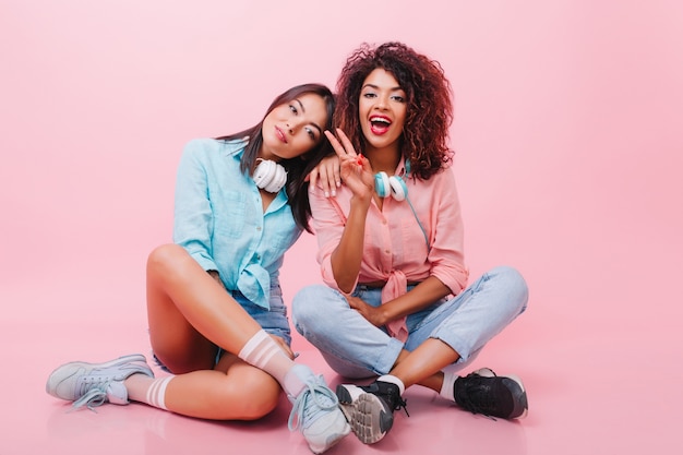 Incrível senhora africana encaracolada sentada no chão perto de romântica menina asiática de camisa azul. foto interna de modelos femininos elegantes posando no chão com as pernas cruzadas e rindo.