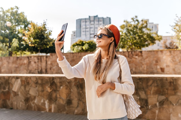 Incrível senhora francesa de camisa branca, fazendo selfie no fim de semana de outono. Adorável garota deslumbrante na boina vermelha em pé na rua com o telefone.