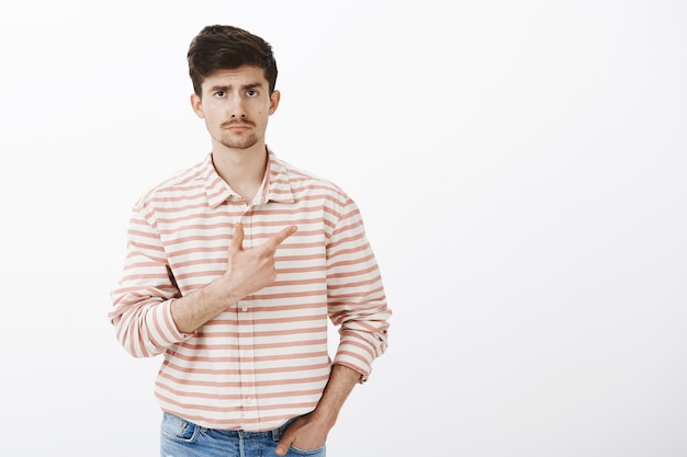 indiferente e desinteressado modelo masculino, caucasiano, com barba e bigode, segurando a mão no bolso e apontando para o canto superior direito com rosto sombrio