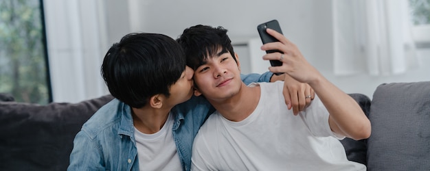Influenciador asiático Casal gay vlog em casa. Os homens asiáticos LGBTQ felizes relaxam o divertimento usando o upload do vídeo do vlog do estilo de vida do registro do telefone móvel da tecnologia nas mídias sociais ao encontrar o sofá na sala de estar.