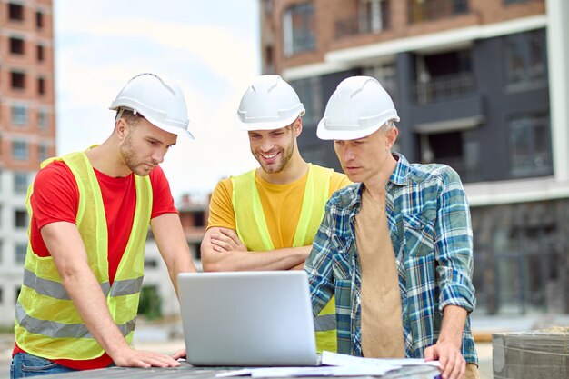 Informação importante. Três homens com capacete protetor olhando para o laptop com interesse enquanto estão em um canteiro de obras durante o dia