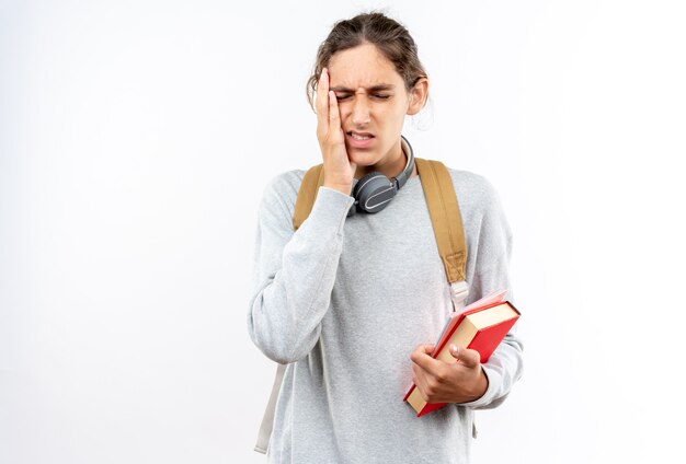 Insatisfeito com os olhos fechados, jovem estudante usando uma mochila com fones de ouvido no pescoço, segurando livros, colocando a mão em um dente dolorido isolado na parede branca