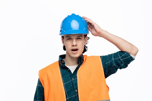Insatisfeito jovem engenheiro masculino usando capacete de segurança e colete de segurança tocando capacete olhando para câmera isolada no fundo branco