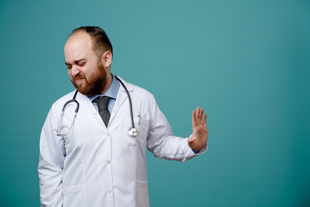 Insatisfeito jovem médico masculino vestindo jaleco e estetoscópio em volta do pescoço, não mostrando nenhum gesto para o lado com os olhos fechados, isolados em fundo azul com espaço de cópia