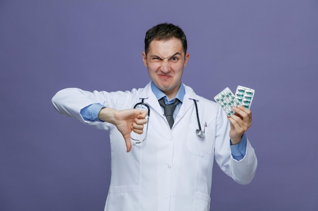 Insatisfeito jovem médico masculino vestindo roupão médico e estetoscópio no pescoço, olhando para a câmera mostrando pacotes de pílulas e polegar para baixo isolado no fundo roxo