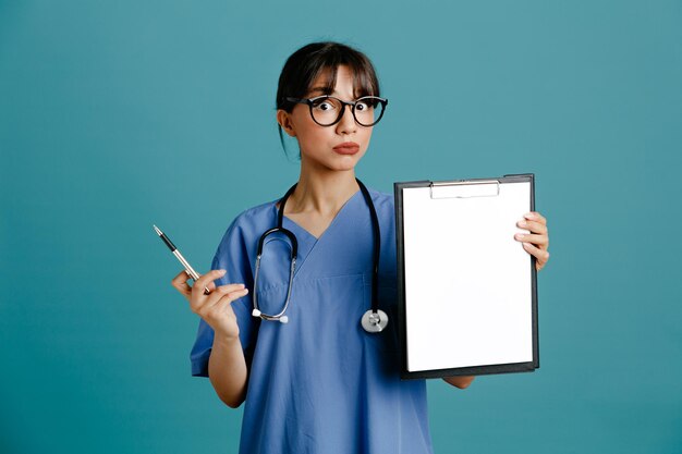 Insatisfeito segurando a prancheta jovem médica usando estetoscópio uniforme de quinto isolado em fundo azul