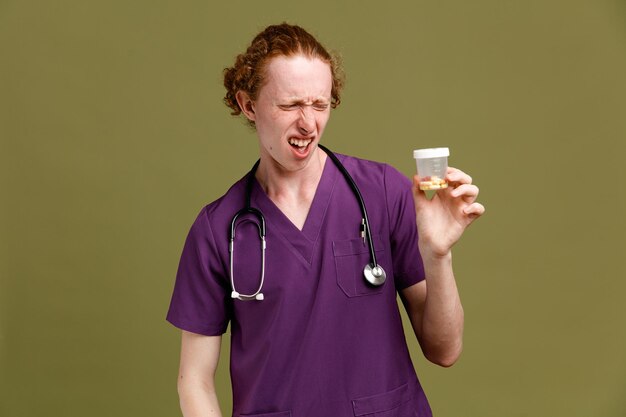Insatisfeito segurando lata de comprimidos jovem médico masculino vestindo uniforme com estetoscópio isolado em fundo verde