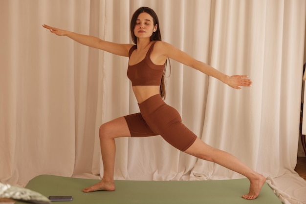 Instrutor de ioga feminino caucasiano jovem magro fazendo pose de pé no tapete em quarto espaçoso com luz do dia Morena no top marrom e legging fechou os olhos conceito de meditação