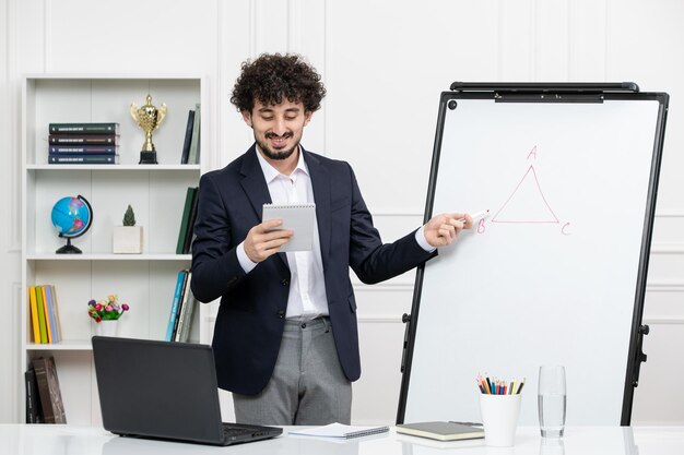 Instrutor moreno professor com computador de terno e quadro branco em sala de aula olhando notas