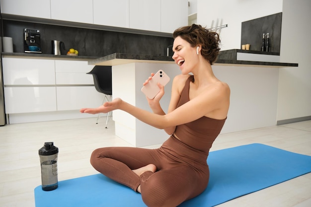 Foto grátis instrutora de exercícios de fitness despreocupada senta-se em casa com tapete de ioga de garrafa de água usa pecado de agasalho