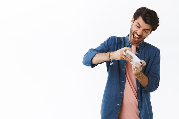 Intenso homem caucasiano bonito jogando intenso jogo legal virando smartphone e inclinando a cabeça como quer ganhar a competição usando fones de ouvido sem fio em pé fundo branco emocionado